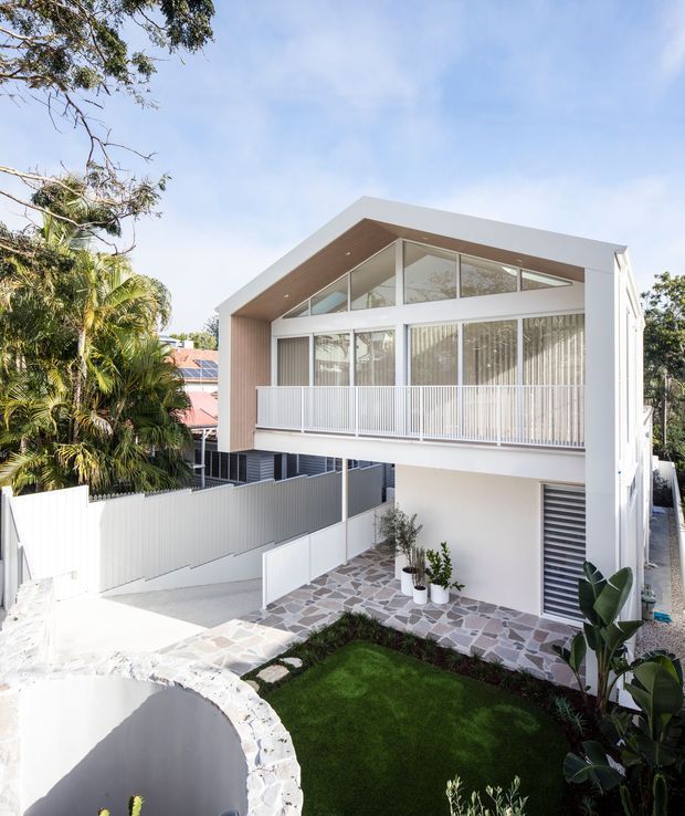 Bright exterior view of a contemporary multi-level residence with a distinctive gabled roof form, white rendered facade, expansive glazing, and a covered upper balcony with vertical balustrading. The property includes a paved courtyard and front garden, surrounded by established tropical vegetation and neighbouring homes.