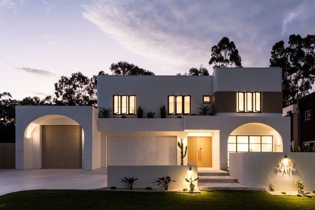 Twilight exterior photograph of a contemporary two-storey residence with a white rendered facade, flat rooflines, arched garage and entry openings, warm illuminated windows, a central garage, landscaped front garden, and a dark neighboring structure partially visible at the side.