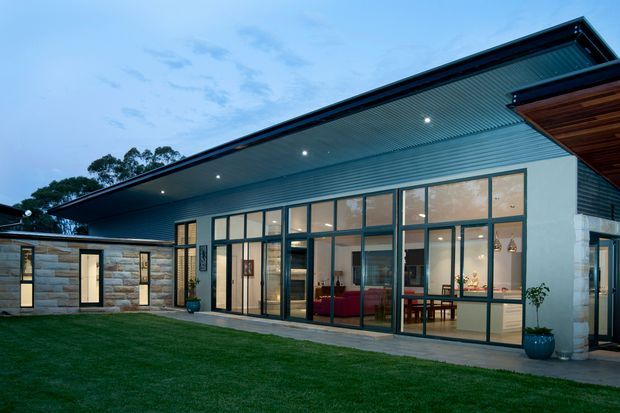 Exterior dusk view of a contemporary single-storey residence with a broad projecting roof canopy, extensive floor-to-ceiling glazed doors and windows, dark metal framing, pale stone-clad walls, and a covered outdoor terrace opening onto a lawn. Warm interior lighting reveals living and dining areas inside.
