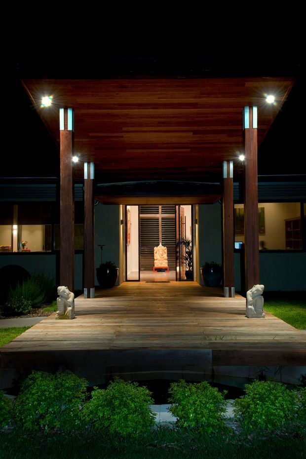 Night-time exterior view of a dramatic entrance pavilion with a broad timber-clad canopy supported by dark timber posts. A timber deck walkway leads to illuminated glass and timber entry doors, with small sculptural figures and landscaped planting at the front. The dark setting and warm architectural lighting emphasize the symmetrical, refined entrance design.