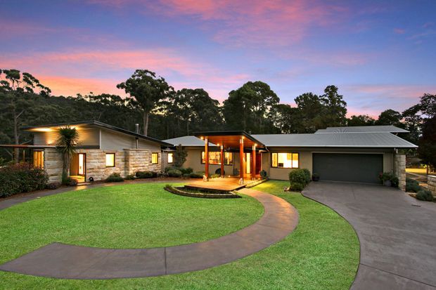Wide dusk exterior view of a low-profile single-storey residence set within a landscaped suburban or bush setting. The home features a long, gently pitched roofline, warm timber-lined entry pavilion, stone and timber-clad walls, illuminated windows, an integrated double garage, curved concrete paths, and a broad manicured lawn.