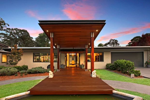 A low-slung single-storey residence viewed from the front at dusk, featuring a broad projecting entry canopy with a warm timber-lined soffit, substantial timber posts, and a raised timber walkway leading to glazed entry doors. The home has horizontal modern rooflines, light rendered walls, dark window frames, manicured lawn and planting, and warm interior lighting, set against mature trees and a colourful evening sky.