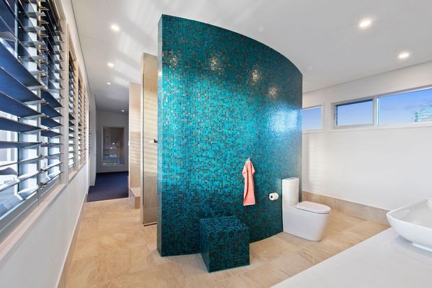 Bright contemporary bathroom with a curved turquoise mosaic-tile feature wall dividing the toilet and vanity areas from the adjoining space. The room has pale stone-look flooring, white sanitaryware, a white vanity, recessed ceiling lights, high horizontal windows, and timber-framed louvred glazing along one side.