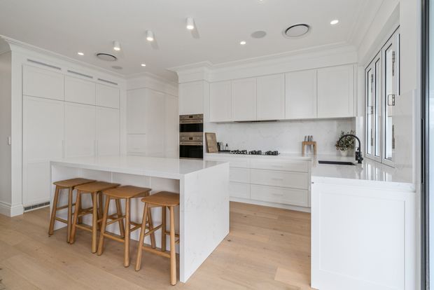 Bright, spacious white kitchen with extensive shaker-style cabinetry, a large waterfall-edge island with four timber stools, integrated appliances, pale timber flooring, and a light stone or marble-look splashback. Recessed and pendant-style ceiling lighting, decorative cornices, and large black-framed windows create a refined contemporary interior.