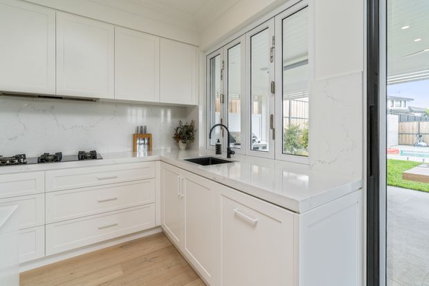 Bright white L-shaped kitchen with shaker-style cabinetry, a pale stone benchtop and matching marble-look splashback. The kitchen includes a gas cooktop, undermount sink, brushed metal tapware, timber-look flooring and extensive windows and glazed doors opening toward an outdoor area.