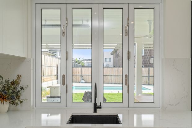 Bright contemporary kitchen interior featuring a large white-framed multi-panel bi-fold window above a dark undermount sink and minimalist black tap. The window overlooks a fenced backyard with a swimming pool. White cabinetry, pale stone benchtops, and subtle grey veining create a clean, light-filled space.