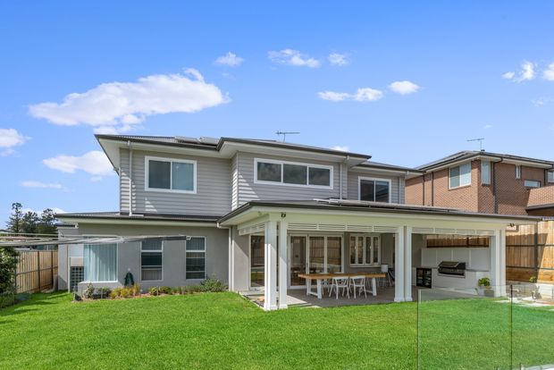 Exterior rear view of a two-storey suburban house with light grey horizontal weatherboard cladding, a low-pitched roof, white-framed windows, and a covered outdoor entertaining area. The covered patio includes a dining table and built-in barbecue, opening onto a broad green lawn enclosed by fencing. Adjacent brick housing is visible in the background.