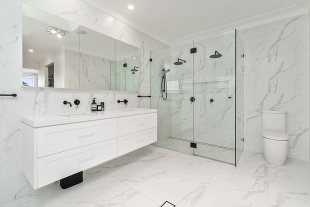 Bright contemporary bathroom featuring full-height white marble-look surfaces with grey veining, a floating white double vanity, large mirrored cabinetry, matte black tapware and fixtures, and a frameless glass walk-in shower with dual shower heads. A white toilet sits against the marble-look wall, with recessed ceiling lighting and subtle decorative ceiling trim.