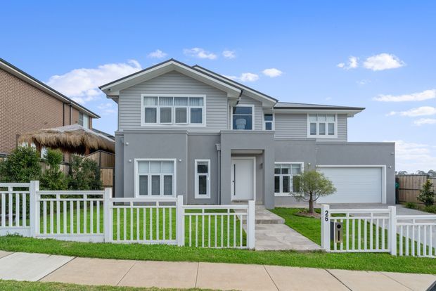 Street-facing view of a contemporary two-storey suburban house with a grey rendered lower facade, light grey horizontal weatherboard cladding to the upper gabled section, white-framed windows, a covered entry, attached double garage, front lawn, and white picket-style fencing. The home has a mixed gable-and-low-pitched roof form and a neat landscaped frontage.