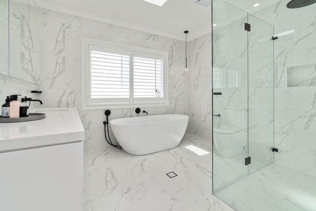 Bright contemporary bathroom featuring full-height white marble-look surfaces with grey veining, a freestanding white bathtub beneath a shuttered window, a minimalist floating vanity, and a frameless glass walk-in shower with recessed shelving.