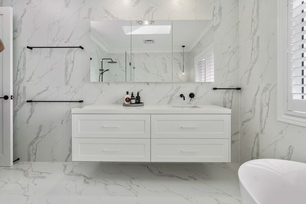 Bright contemporary bathroom with a floating white double vanity, integrated basin, large wall mirror, frameless glass shower enclosure, black tapware and grab rails, and full-height white marble-look veined wall and floor surfaces. A shuttered window and freestanding bathtub are visible at the right.