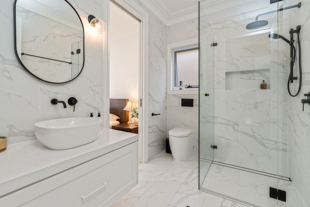 Bright, refined bathroom with white marble-look wall and floor surfaces, a floating white vanity with vessel basin, black tapware, rounded black-framed mirror, concealed toilet, and frameless glass walk-in shower with black fixtures. An open doorway reveals part of an adjacent bedroom and bedside lamp.