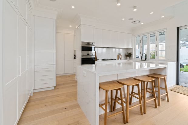 Bright, spacious white kitchen with extensive Shaker-style cabinetry, integrated appliances, a large white island with waterfall edge and seating for four, pale timber flooring, and generous glazed doors and windows opening to the outdoors.