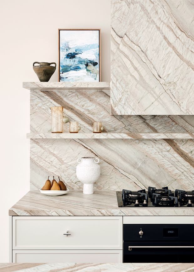 A refined kitchen vignette featuring dramatic veined natural stone wrapping the splashback, open shelving, raised feature panels and benchtops. Pale cabinetry, a black built-in oven and black gas cooktop create contrast, while ceramics, artwork and a bowl of pears add styling detail.
