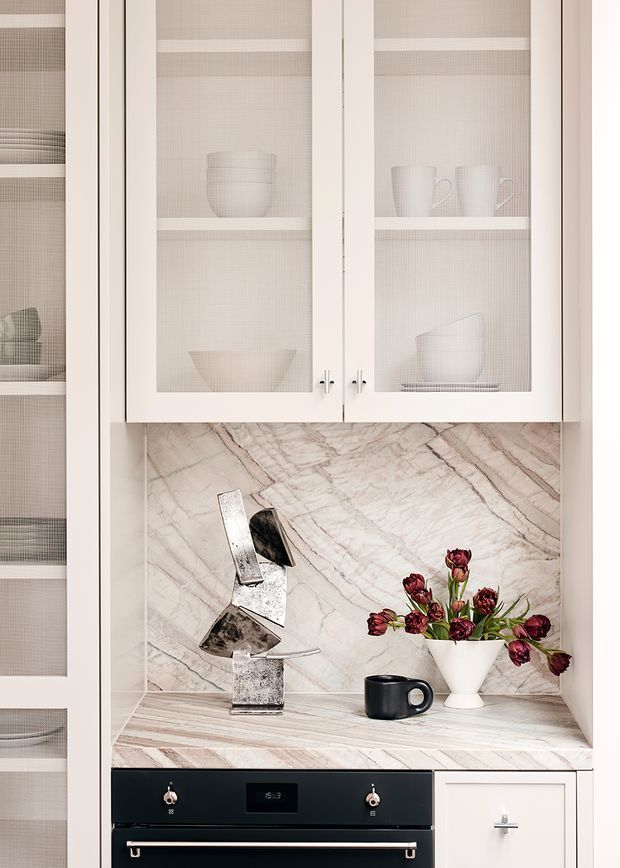 Close-up of a refined kitchen or butler’s pantry wall featuring white framed glass-front cabinetry with fine mesh inserts, displayed white crockery, a pale veined stone splashback and benchtop, and integrated dark oven cabinetry below. Small decorative objects and flowers sit on the counter.