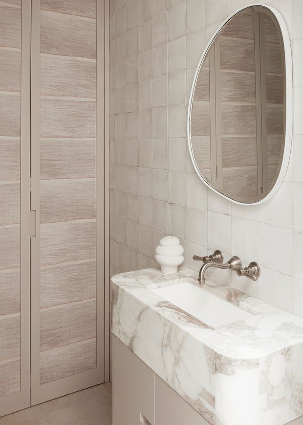A refined bathroom vanity area with a thick white marble benchtop and integrated basin, wall-mounted brushed metal tapware, an oval framed mirror, and pale beige square wall tiles. A tall pale timber-grain built-in cabinet is visible to the left. The palette is soft, warm, and neutral with subtle metallic accents.