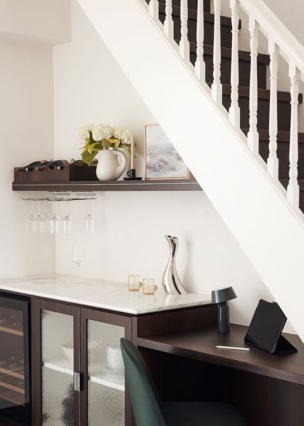 A refined home bar and compact study nook fitted beneath a staircase. Dark timber cabinetry and shelving contrast with a white marble-look benchtop, while hanging stemware, a wine rack, decorative ceramics and a tablet workstation define the space. The staircase has traditional white turned balusters and a dark timber stair finish.