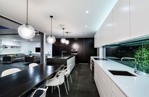 A spacious contemporary kitchen and dining area with sleek white and dark cabinetry, a large central island, integrated appliances, and a long white benchtop beneath a glossy black splashback. A black dining table with white chairs sits in the foreground, while a modern living room is visible through an open connection at the rear. Globe pendant lights and a green planted feature visible behind glazing add visual interest.
