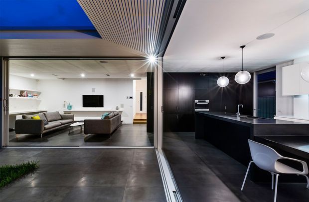 Contemporary open-plan kitchen and living area viewed through large glazed sliding doors. The space features a dark, minimalist kitchen with a long island and integrated cabinetry, white globe pendant lights, dark tiled flooring, and a bright adjoining living room with neutral sofas and built-in shelving. A dramatic slatted timber ceiling feature extends over the transition between the rooms, with strong indoor-outdoor visual continuity.