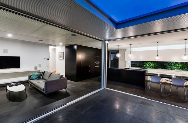 A contemporary open-plan living and kitchen area connected by fully glazed sliding doors to a covered outdoor courtyard or terrace. The interior features dark tiled flooring, a grey sofa and black feature joinery, while the kitchen has a dark island, white cabinetry, pendant lighting and a narrow garden view. A large blue-lit overhead opening or skylight creates a strong architectural focal point.