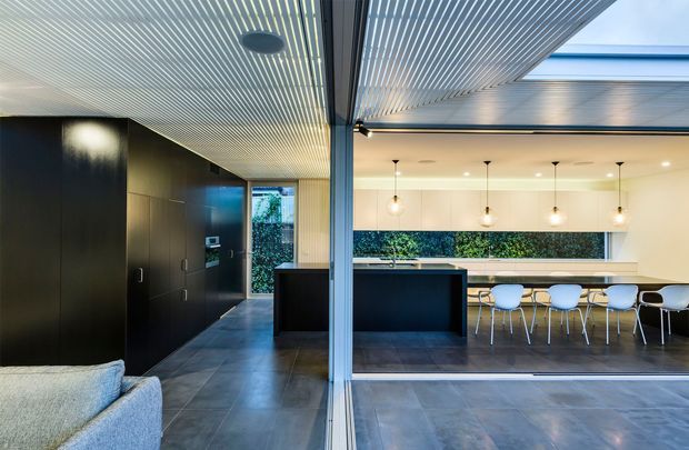 Contemporary open-plan interior viewed through a large glazed opening, combining a dark minimalist kitchen with a dining area. The space features charcoal built-in cabinetry, a central dark island, large-format grey floor tiles, a linear slatted ceiling, a skylight, extensive glazing, and a long dining table with white chairs and pendant lighting. A partially visible sofa suggests an adjoining living area.