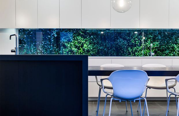 Contemporary kitchen and dining area with a dark island, white flat-panel cabinetry, integrated sinks and taps, and a long horizontal mirrored or glazed backsplash reflecting lush greenery. A dining table with sculptural blue chairs sits behind the island, while a spherical pendant light hangs above. The space has a clean, high-contrast, polished appearance.