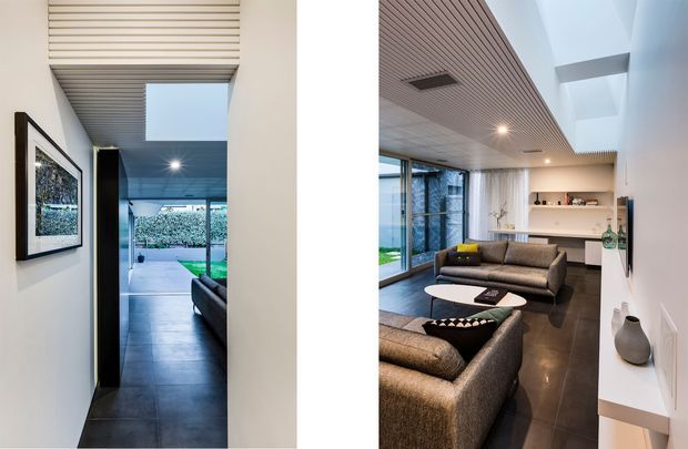 A modern open-plan living room shown from two angles, with dark large-format floor tiles, charcoal upholstered sofas, floor-to-ceiling glazing, white walls, and a timber-look slatted ceiling. A skylight, recessed lighting, built-in shelving, and a slim white console contribute to the clean contemporary interior. The room opens toward a landscaped outdoor area visible through the glazing.