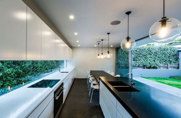 A sleek contemporary galley kitchen with extensive white handleless cabinetry along the left wall, a long dark island with integrated sinks and seating, dark flooring, and a row of globe pendant lights. Full-height glazing and an open connection to a landscaped courtyard bring greenery and daylight into the space.