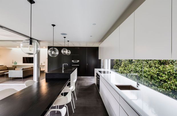 A contemporary open-plan kitchen with a long dark island and integrated dining bench, white handleless cabinetry, dark tiled flooring, and a glossy white perimeter benchtop. Clear globe pendant lights hang above the island, while a full-height window beside the sink overlooks dense greenery. A living area is visible beyond the kitchen.
