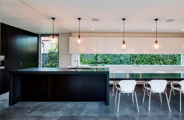 A contemporary open-plan kitchen and dining area with a large dark island, integrated sink and tap, streamlined dark cabinetry, a long dining table with white chairs, pendant lighting, and full-width windows overlooking greenery. The space has a dark tiled floor, white cabinetry and walls, and a linear textured ceiling.