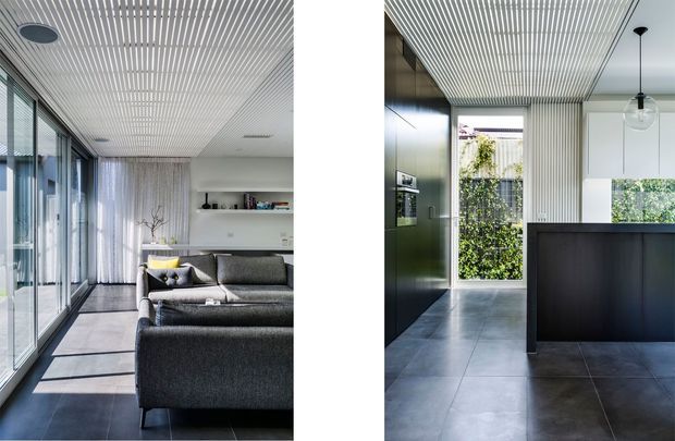 A split view of a bright contemporary home interior, showing an open-plan living area and adjacent kitchen. The living room has full-height glazed doors, a dark grey sectional sofa, sheer curtains, built-in white shelving, and a linear slatted ceiling. The kitchen features dark cabinetry, a central island or peninsula, large-format dark tiled flooring, a glass pendant light, and views toward a planted courtyard through glazing.