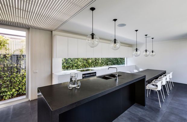 Contemporary kitchen dominated by a large black island with an integrated sink and dark benchtop, white flat-panel cabinetry, and a long adjoining dining counter with white bar stools. Multiple clear-glass pendant lights hang above the island and dining area. Full-height glazing and a horizontal garden outlook bring greenery into the bright, minimalist space. A distinctive timber slatted ceiling and matching vertical wall screen add texture.