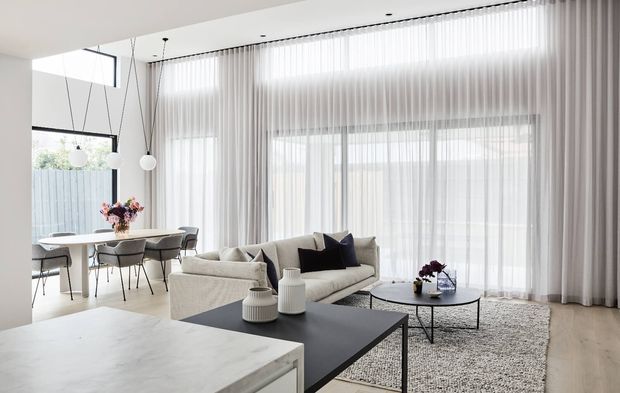 Bright, open-plan contemporary living and dining area with floor-to-ceiling sheer curtains and glazing. A light upholstered sofa, black rectangular coffee table, textured neutral rug, round side table, and sculptural pendant lights create a refined minimalist interior. A dining table and chairs sit behind the living area, while a pale stone-topped kitchen island is visible in the foreground.