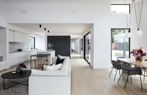 Bright open-plan residential interior combining a minimalist white kitchen, central island, living area, and dining space. Pale timber flooring, white walls and cabinetry, dark built-in kitchen joinery, black-framed glazing, and pendant globe lighting create a clean contemporary scheme with abundant natural light.