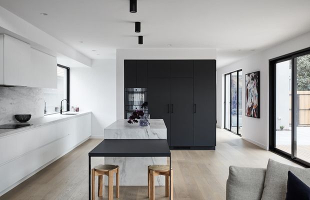 A bright, contemporary open-plan kitchen and living area with white marble-look cabinetry and benchtops, a dark central island and full-height pantry cabinetry, pale timber flooring, black-framed glazing, and minimalist black track lighting. Two timber stools sit at the island, while part of a grey sofa is visible in the foreground.