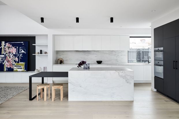 Bright open-plan kitchen with a large white marble-look island, white cabinetry, dark full-height pantry cabinetry, integrated appliances, a black dining table with timber stools, and a marble-look splashback. Contemporary artwork and black track-style ceiling lights add contrast.
