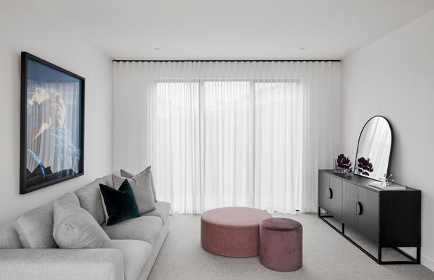 A bright, refined contemporary living room with a large pale sectional sofa, sheer full-height curtains, a black-framed sideboard, oval mirror, framed artwork, and two rounded dusty-rose ottomans. The room has a soft neutral palette with a dark teal cushion and abundant diffused daylight.