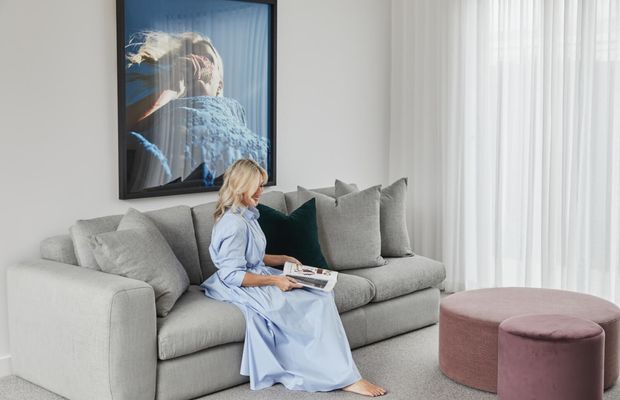 Bright, softly styled living room with a large light-grey sectional sofa, layered grey and deep teal cushions, and two blush-pink upholstered round ottomans. A woman in a pale blue dress is seated reading, with a large framed artwork above the sofa and sheer floor-to-ceiling curtains filtering daylight. The room has a calm, refined contemporary feel.