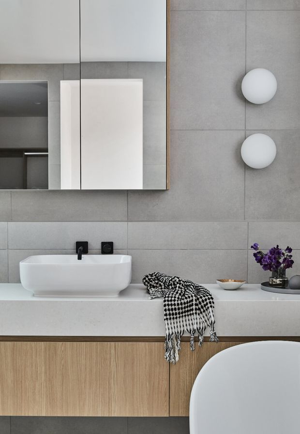 A contemporary bathroom vanity with a white vessel basin, matte black wall-mounted tapware, large mirror, grey tiled walls, and light timber cabinetry. Minimal spherical wall lights and a small decorative arrangement add restrained detail, while a patterned monochrome textile drapes over the vanity.