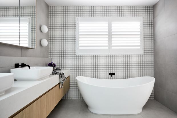 Bright contemporary bathroom featuring a freestanding white bathtub, double white vessel basins on a timber vanity, grey tiled walls and floor, a small square mosaic-tile feature wall, white plantation shutters, and a large mirror.