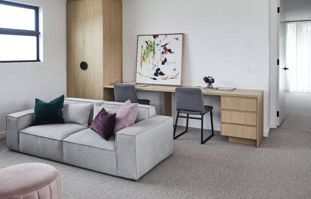 A bright, contemporary living room with a large light-grey modular sofa, teal and purple cushions, and a pale pink upholstered ottoman. A timber built-in wall unit incorporates a tall cupboard and a long desk with drawers, paired with a grey chair. Neutral carpet, white walls, a black-framed window, and abstract artwork create a calm, refined interior. An adjoining carpeted room is visible through the doorway.