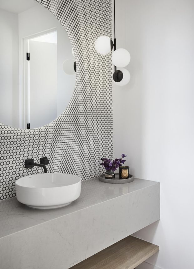 Contemporary bathroom vanity with a white vessel basin on a pale stone benchtop, matte black wall-mounted tapware, and a floating vanity with an open timber shelf. The feature wall is clad in small round black-and-white mosaic tiles and paired with a large rounded mirror. A sculptural black-and-white pendant light and minimal accessories complete the refined monochromatic scheme.