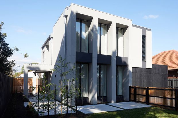 Exterior view of a contemporary two-storey residence with a crisp white rendered facade, strong vertical window bays, dark window surrounds and screening, and a flat roofline. A paved path, small lawn, timber fencing, and a covered entry structure frame the front elevation.