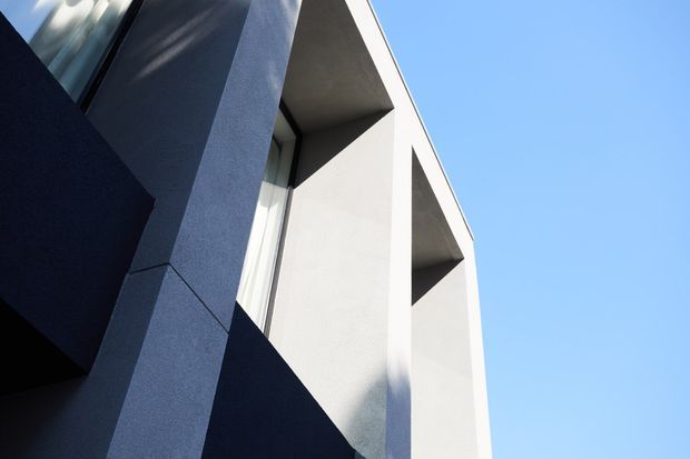 Upward-looking close view of a modern angular building facade with smooth pale rendered walls, deep recessed window openings, dark blue-grey projecting planes, and sharp geometric lines against a clear blue sky.