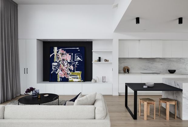 A bright open-plan contemporary living, dining and kitchen interior. The room features white built-in cabinetry, a large abstract artwork set within dark vertical wall panelling, a pale sectional sofa, black round coffee tables, a black dining table with timber stools, light timber flooring, and a pale marble-look kitchen island and splashback. Full-height grey curtains and restrained black accents create a refined monochromatic palette.