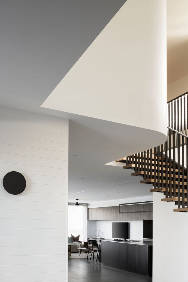 A contemporary open-plan interior featuring a dramatic sculptural curved white ceiling and soffit, a floating staircase with dark vertical metal balusters and timber treads, and a living area with dark built-in cabinetry, television, and furniture. The walls are predominantly white with horizontal wall panelling, complemented by dark grey flooring and charcoal joinery.
