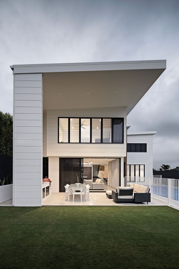 Contemporary two-storey residential home viewed from the rear garden, with a large cantilevered flat roof and deep covered outdoor entertaining area. The facade combines white horizontal cladding with dark-framed glazing. A paved patio includes an outdoor dining setting, barbecue cabinetry and lounge seating, opening directly into a bright living area. A white balustrade encloses the upper-level terrace or side edge, with manicured lawn in the foreground.