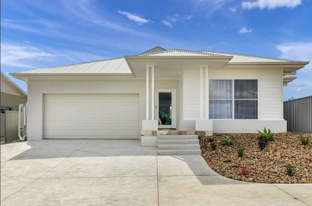 Single-storey suburban home viewed from the front, with a pale rendered and weatherboard-style facade, a low-pitched light metal hipped roof, covered entry porch with columns, large front window, attached single garage, concrete driveway, and a landscaped rock garden.