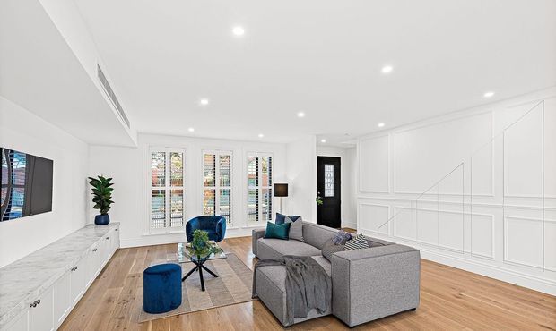 Bright contemporary living room with a grey sectional sofa, blue accent ottoman, small coffee table, wall-mounted television, built-in white media cabinetry, white decorative wall panelling, timber flooring, plantation shutters, and recessed ceiling lighting.