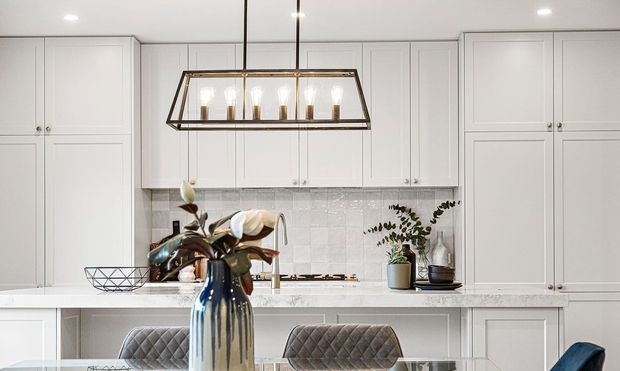 Bright, refined kitchen with extensive white shaker-style cabinetry, a pale stone island benchtop, glossy textured white tile splashback, and a contemporary rectangular pendant with bronze or dark metal framing. A reflective ceramic vase and upholstered bar stools add subtle contrast in the foreground.